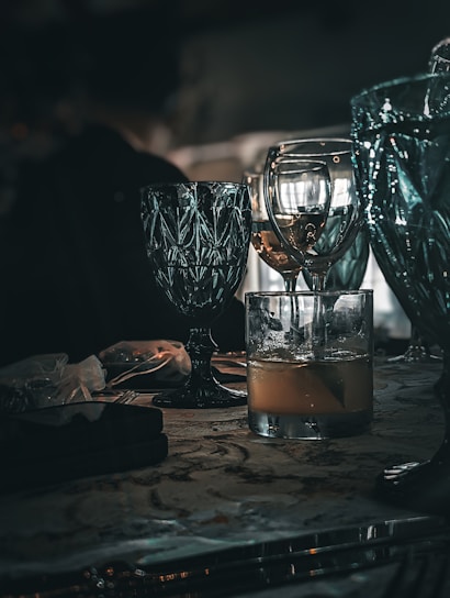 A collection of elegant glassware is placed on a table, featuring a mix of turquoise and clear glasses. The scene is dimly lit, creating reflections and shadows that enhance the luxurious feel. The table surface has a patterned design and holds additional items, including a small black object and a translucent bag.
