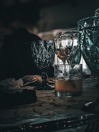 A collection of elegant glassware is placed on a table, featuring a mix of turquoise and clear glasses. The scene is dimly lit, creating reflections and shadows that enhance the luxurious feel. The table surface has a patterned design and holds additional items, including a small black object and a translucent bag.