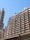 A cluster of high-rise residential buildings with numerous windows and balconies under a clear blue sky. The architectural style is modern, and the structures exhibit a uniform, grid-like appearance. A part of an overhead structure is visible at the bottom of the frame.