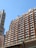 A cluster of high-rise residential buildings with numerous windows and balconies under a clear blue sky. The architectural style is modern, and the structures exhibit a uniform, grid-like appearance. A part of an overhead structure is visible at the bottom of the frame.
