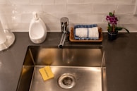 A modern kitchen sink with a chrome faucet is set into a dark countertop. Nearby, there is a yellow sponge labeled 'CALDERA', a soap dispenser, tissues on a towel, and a small potted plant with purple flowers.