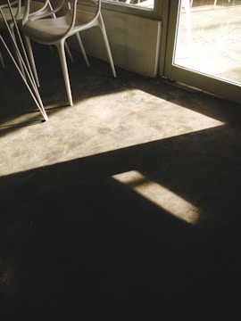 Sunlight creates geometric patterns with light and shadow on a concrete floor, highlighting a chair and a window frame. The subtle play of light evokes a sense of stillness and simplicity within an industrial interior setting.
