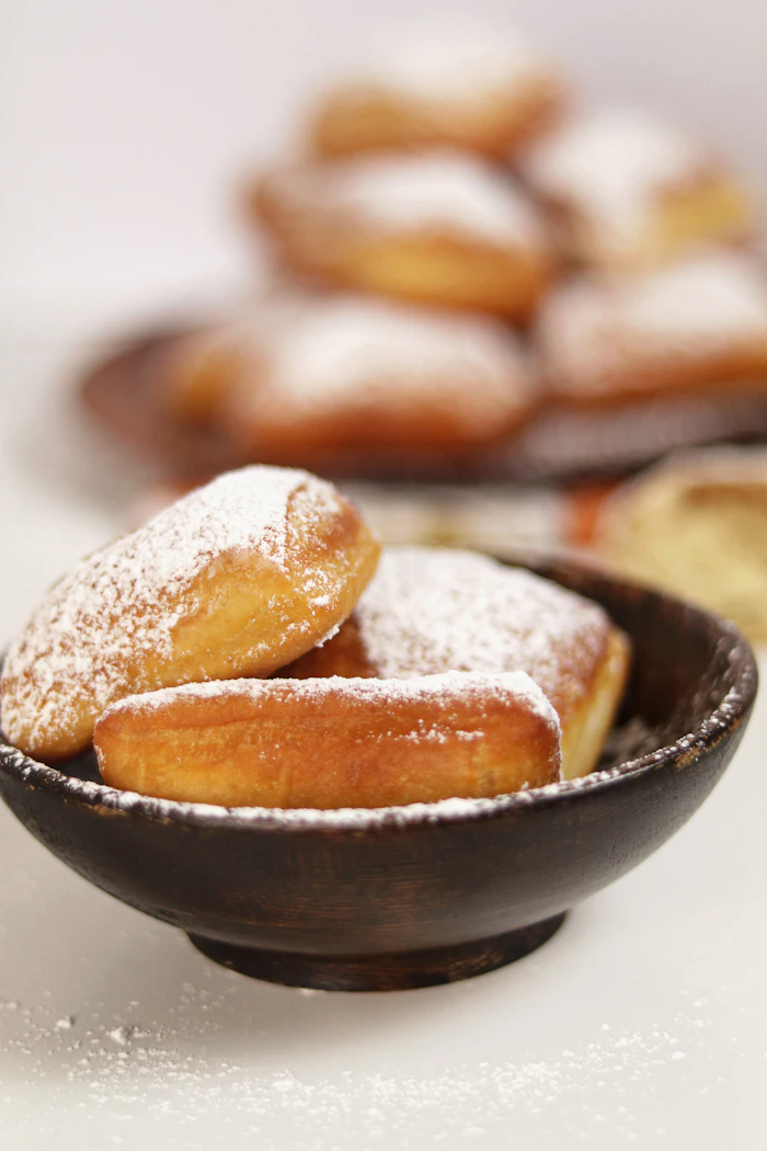 Beignets heaped in a dark wooden bowl with powdered sugar scattered around