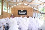 A large group of people wearing white hijabs is gathered in an outdoor but roofed wooden structure. They are facing a stage with a black banner that reads 'Halal Bihalal SMK Ulil Albab NW.' Several speakers are seated behind a table on the stage.