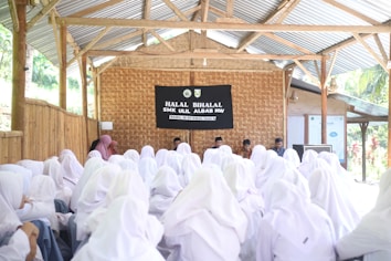 A large group of people wearing white hijabs is gathered in an outdoor but roofed wooden structure. They are facing a stage with a black banner that reads 'Halal Bihalal SMK Ulil Albab NW.' Several speakers are seated behind a table on the stage.