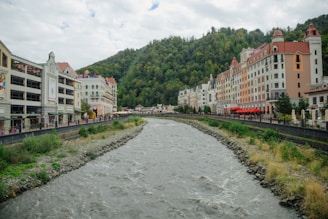 A river flows through a picturesque town flanked by colorful buildings with European architectural styles. The buildings have orange and peach facades with red roofs, and some feature café umbrellas. Lush, green hills rise in the background under a cloudy sky.
