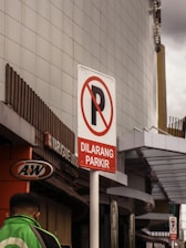 A parking prohibition sign with the text 'Dilarang Parkir' set against an urban background. The sign is prominently displayed on a pole, with a sleek, modern building in the backdrop. A person in a green jacket is visible, walking on the left side near an A&W restaurant sign.