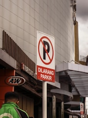A parking prohibition sign with the text 'Dilarang Parkir' set against an urban background. The sign is prominently displayed on a pole, with a sleek, modern building in the backdrop. A person in a green jacket is visible, walking on the left side near an A&W restaurant sign.