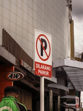 A parking prohibition sign with the text 'Dilarang Parkir' set against an urban background. The sign is prominently displayed on a pole, with a sleek, modern building in the backdrop. A person in a green jacket is visible, walking on the left side near an A&W restaurant sign.