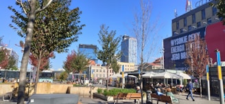 A bustling urban scene with people sitting at outdoor cafes, trees lining the pavements, modern high-rise buildings in the background, and a large billboard on a building. The sky is clear and blue, creating a vibrant atmosphere.