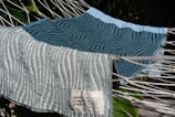 Two textured towels with wavy patterns are laid over a hammock. The towel in the foreground is light gray with darker gray wavy lines, and the one in the background is teal with similar patterns. A tag with text is visible on the light gray towel. The hammock ropes are white and there are green leaves in the background.