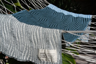Two textured towels with wavy patterns are laid over a hammock. The towel in the foreground is light gray with darker gray wavy lines, and the one in the background is teal with similar patterns. A tag with text is visible on the light gray towel. The hammock ropes are white and there are green leaves in the background.