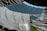 Two textured towels with wavy patterns are laid over a hammock. The towel in the foreground is light gray with darker gray wavy lines, and the one in the background is teal with similar patterns. A tag with text is visible on the light gray towel. The hammock ropes are white and there are green leaves in the background.