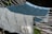 Two textured towels with wavy patterns are laid over a hammock. The towel in the foreground is light gray with darker gray wavy lines, and the one in the background is teal with similar patterns. A tag with text is visible on the light gray towel. The hammock ropes are white and there are green leaves in the background.