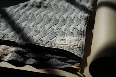 A folded textile piece made from 100% organic cotton with a geometric chevron pattern. The label, indicating it's from towel studio and made in Germany, is visible. The fabric appears to be draped on a flat surface, with one edge slightly in shadow.
