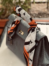 A luxurious handbag with an ostrich leather pattern adorned with a colorful, branded silk scarf wrapped around the handle. The scarf features a design with black and white patterns accented by bold orange stripes. The background is blurred, suggesting an outdoor setting with greenery.