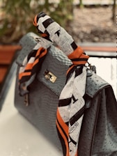 A luxurious handbag with an ostrich leather pattern adorned with a colorful, branded silk scarf wrapped around the handle. The scarf features a design with black and white patterns accented by bold orange stripes. The background is blurred, suggesting an outdoor setting with greenery.