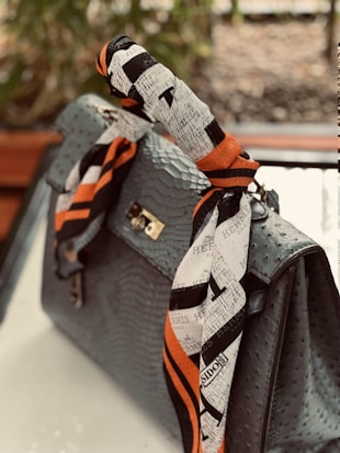 A luxurious handbag with an ostrich leather pattern adorned with a colorful, branded silk scarf wrapped around the handle. The scarf features a design with black and white patterns accented by bold orange stripes. The background is blurred, suggesting an outdoor setting with greenery.