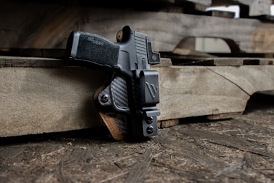 A handgun is securely fastened in a black holster, resting against a stack of wooden pallets. The setting appears rustic with wooden textures in the background, and the gun's grip features a textured pattern for enhanced grip.