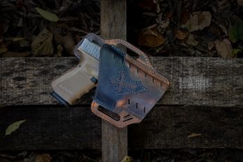 A tan handgun rests in a black leather holster, placed on a rustic wooden surface surrounded by dry leaves.