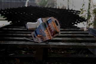 A handgun is placed inside a brown leather holster featuring a star design. The holster rests on a wooden surface with a textured metal background blurred in the distance.