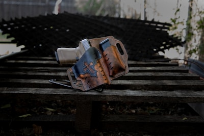 A handgun is placed inside a brown leather holster featuring a star design. The holster rests on a wooden surface with a textured metal background blurred in the distance.