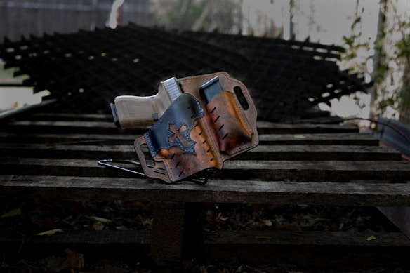 A handgun is placed inside a brown leather holster featuring a star design. The holster rests on a wooden surface with a textured metal background blurred in the distance.