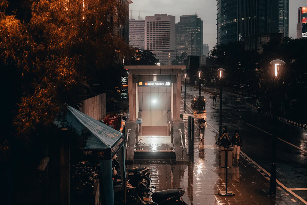 Rain-darkened pavement around the Dukuh Atas BNI metro entrance beneath Jakarta office towers at dusk