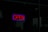 A brightly lit neon sign displaying the word 'OPEN' in red, surrounded by a purple border, hangs in what appears to be a storefront window. The surrounding area is dimly lit, suggesting evening or nighttime.