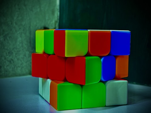 A partially solved Rubik's cube showing various colored squares. The colors visible include green, red, blue, white, and orange. The cube is placed on a smooth surface with a dark background.