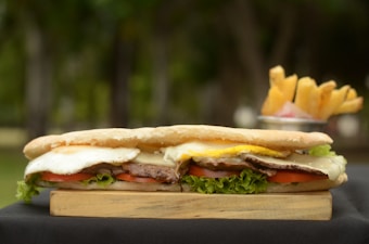A long sandwich filled with layers of ingredients such as lettuce, tomato slices, fried egg, meat, and cheese is placed on a wooden board. In the background, a metal container holds a portion of golden, crispy fries.