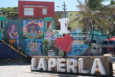 A vibrant urban scene features colorful street art and murals on building walls, depicting various characters and abstract designs. A prominent installation of the phrase 'I heart La Perla' stands in front, with a large white letter 'I' and a red heart replacing the word 'love'. A nearby palm tree suggests a tropical setting, further emphasized by the bright sunlight. Buildings in the background showcase different colors, including red and yellow facades.