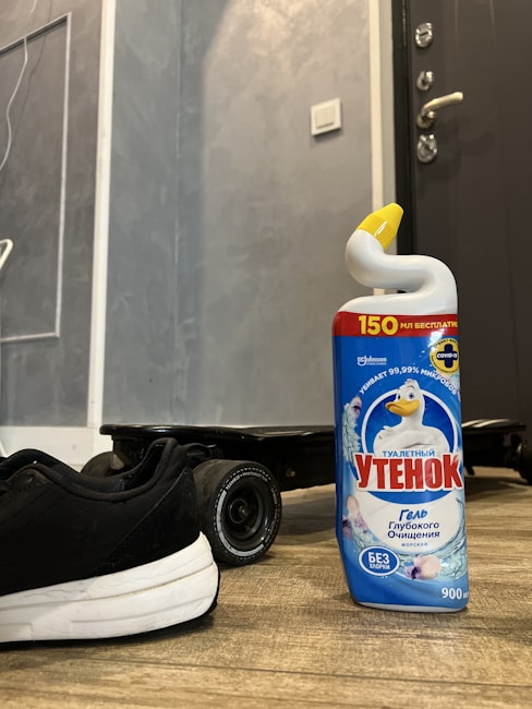 A bottle of cleaning gel with a yellow cap stands on a wooden floor next to a black sneaker and the wheel of a skateboard. The background features a gray textured wall and part of a door with a handle.