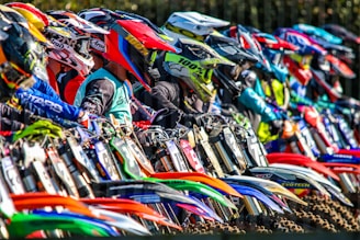 A group of motocross riders lined up, each wearing colorful helmets and gear. Their motorcycles are vibrant and varied in design, highlighting the diversity of the sport. The scene is dynamic, full of energy and anticipation.