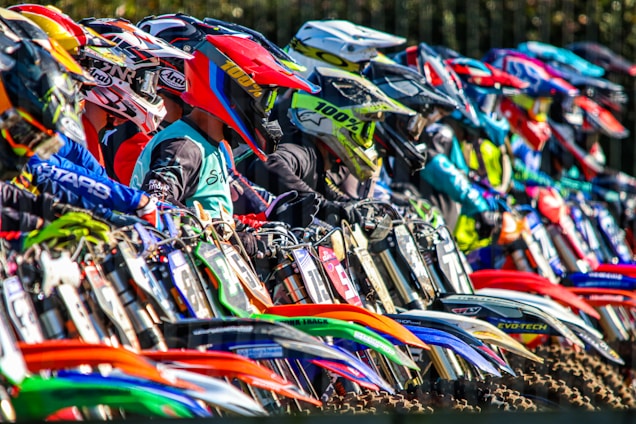 A group of diverse motocross riders smiling together on their bikes at a sunny outdoor track.