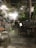 A cozy cafe interior with a rustic and natural ambiance. There are several tables and chairs, some occupied by people engaged in conversation. The decor features a mix of greenery, including large potted plants and hanging vines. Overhead, warm lights illuminate the scene, creating a welcoming atmosphere.