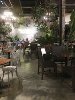 A cozy cafe interior with a rustic and natural ambiance. There are several tables and chairs, some occupied by people engaged in conversation. The decor features a mix of greenery, including large potted plants and hanging vines. Overhead, warm lights illuminate the scene, creating a welcoming atmosphere.