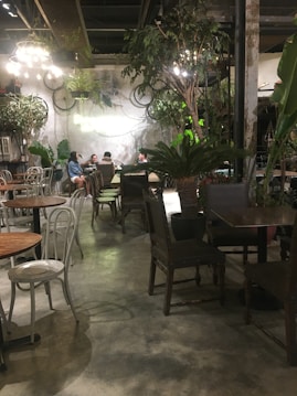 A cozy cafe interior with a rustic and natural ambiance. There are several tables and chairs, some occupied by people engaged in conversation. The decor features a mix of greenery, including large potted plants and hanging vines. Overhead, warm lights illuminate the scene, creating a welcoming atmosphere.