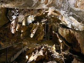 A large cave with rugged rock formations and stalactites hanging from the ceiling. The cave is illuminated by artificial lighting that highlights the various textures and colors of the rocks. Stairs and railings are installed to guide visitors through the cave.