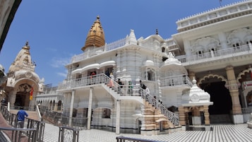 A large, intricately detailed white temple with golden domes rises against a clear blue sky. Several people are walking up the staircase leading to the temple entrance. The architecture features ornate carvings, arches, and a mixture of rounded and angular structures.