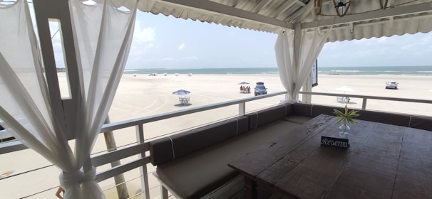 A beach scene viewed from a shaded terrace, featuring white curtains and a rustic wooden table with a 'Reserva' sign. The soft blue sky and ocean meet at the horizon, with scattered clouds overhead. Beach chairs shaded by umbrellas and a few vehicles are visible on the sandy expanse.