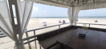 A beach scene viewed from a shaded terrace, featuring white curtains and a rustic wooden table with a 'Reserva' sign. The soft blue sky and ocean meet at the horizon, with scattered clouds overhead. Beach chairs shaded by umbrellas and a few vehicles are visible on the sandy expanse.