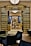 An elegant interior featuring a luxurious hallway. The space is adorned with gold and dark accents, with tall curtains and polished floors. Plants add a touch of greenery, and modern seating surrounds a glass-topped table in the foreground. A central round table, partially obscured, displays decorative items, while ambient lighting complements the sophisticated atmosphere.