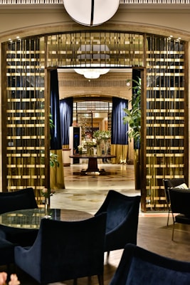 An elegant interior featuring a luxurious hallway. The space is adorned with gold and dark accents, with tall curtains and polished floors. Plants add a touch of greenery, and modern seating surrounds a glass-topped table in the foreground. A central round table, partially obscured, displays decorative items, while ambient lighting complements the sophisticated atmosphere.