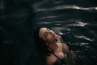A woman is partially submerged in dark, rippling water with her eyes closed and head tilted slightly back. The lighting creates a serene, almost ethereal atmosphere.