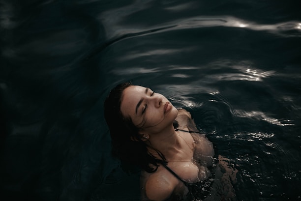 A woman is partially submerged in dark, rippling water with her eyes closed and head tilted slightly back. The lighting creates a serene, almost ethereal atmosphere.
