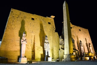 An ancient Egyptian temple facade with large stone statues of pharaohs and an obelisk featuring hieroglyphs, illuminated at night against a dark sky.