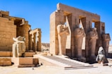 Ancient Egyptian ruins featuring a row of large, carved stone statues resembling pharaohs with crossed arms. Intricate hieroglyphics and detailed carvings decorate the walls and pillars. Bright blue sky and the arid landscape add to the scene.