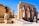 Ancient Egyptian ruins featuring a row of large, carved stone statues resembling pharaohs with crossed arms. Intricate hieroglyphics and detailed carvings decorate the walls and pillars. Bright blue sky and the arid landscape add to the scene.