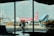 A red and white AirAsia airplane is parked at an airport gate. A worker in a safety vest is cycling in the foreground, silhouetted against the large glass windows of an airport terminal. The scene captures a busy airport environment with ground support equipment and a boarding stair vehicle nearby.